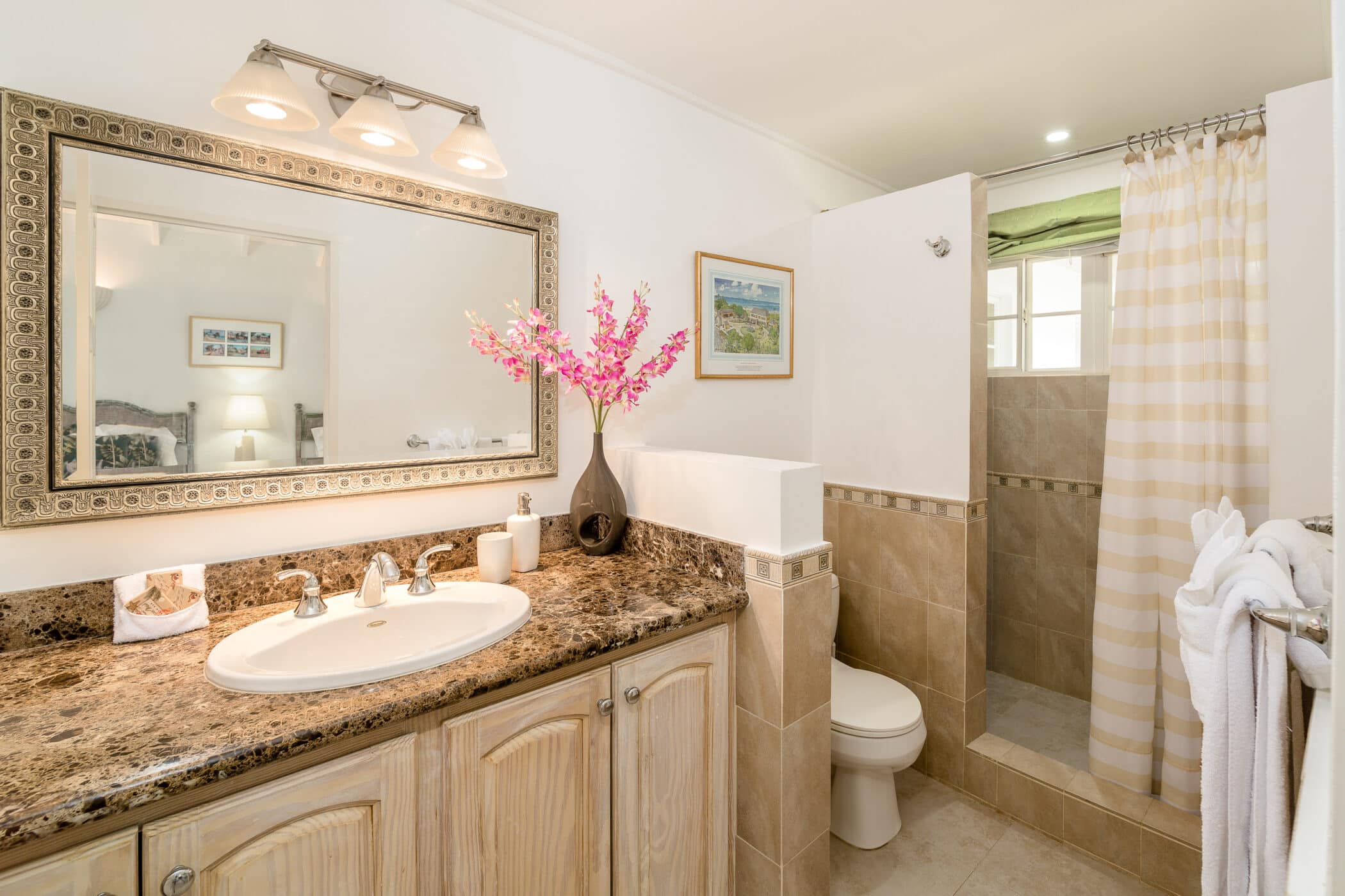 A bathroom with a granite countertop, sink, mirror, wood cabinets, and pink Rose of Sharon flowers in a vase adds a charming touch. There’s a toilet and a tiled shower with a beige striped curtain, while towels hang neatly on a rack near the shower.