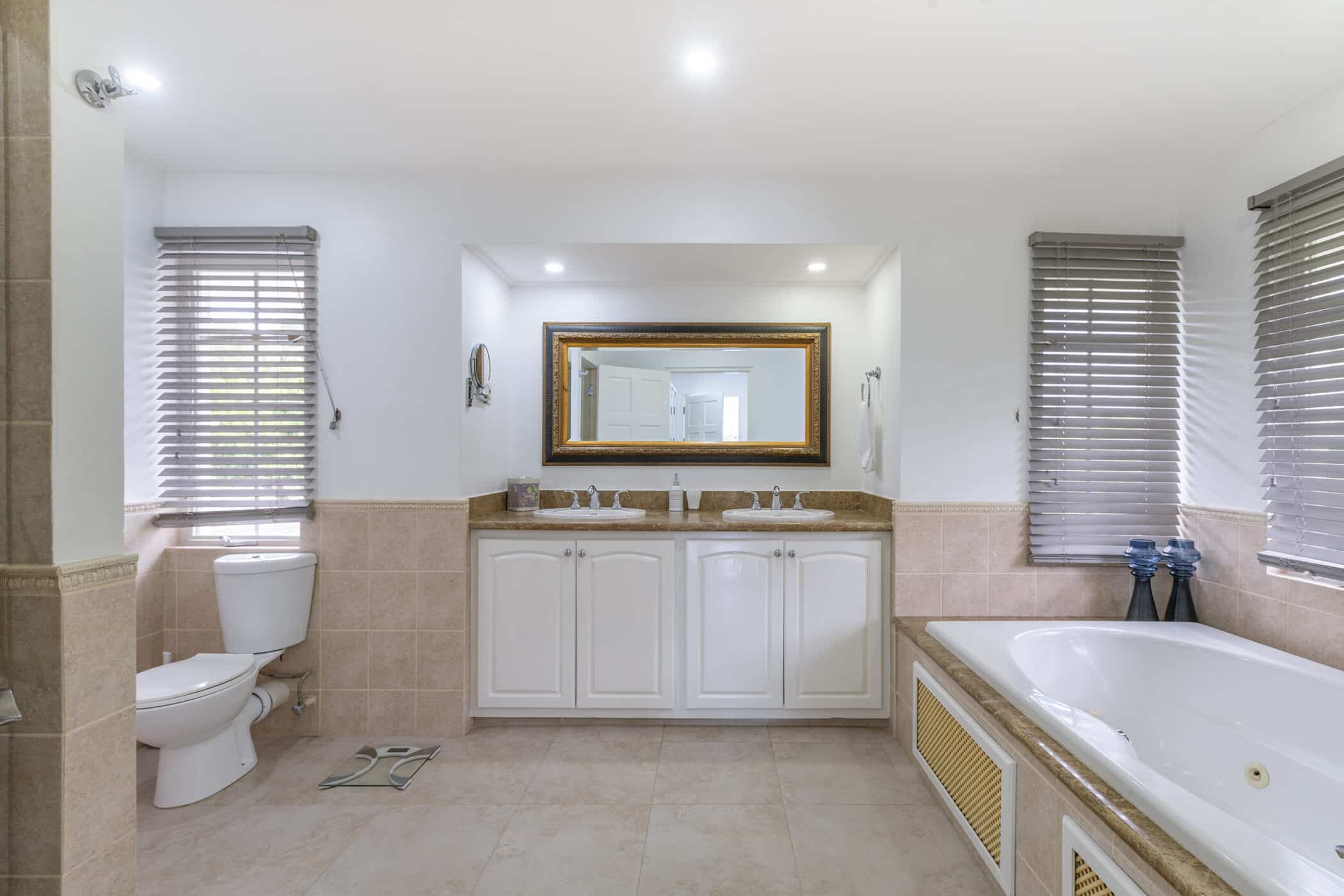 Spacious bathroom with a double sink vanity, large mirror, bathtub, toilet, tiled floor and walls, and two windows with gray blinds. Decorative vases sit by the tub, featuring elegant Rose of Sharon arrangements that add a touch of natural beauty to the serene space.
