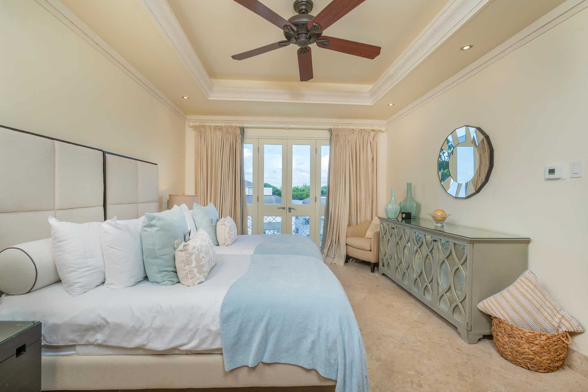 A bright bedroom at Smugglers Cove Penthouse features two beds with light blue and cream bedding, a ceiling fan, a mirrored dresser adorned with vases, a round mirror, and large windows with sheer curtains letting in natural light.
