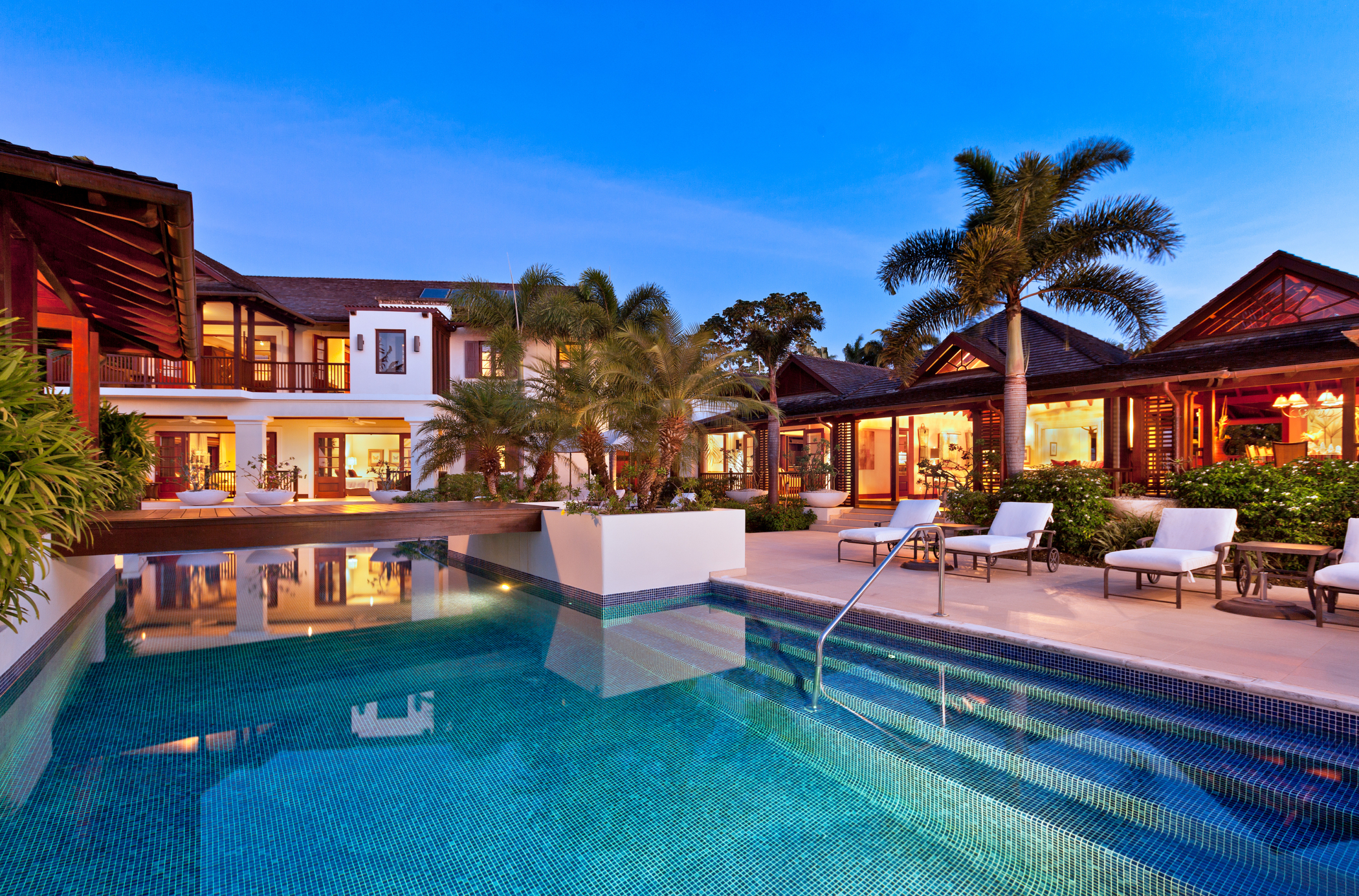 Luxury Alila villa with glowing lights, large swimming pool, palm trees, and lounge chairs at sunset. The modern house features glass doors and tropical landscaping.