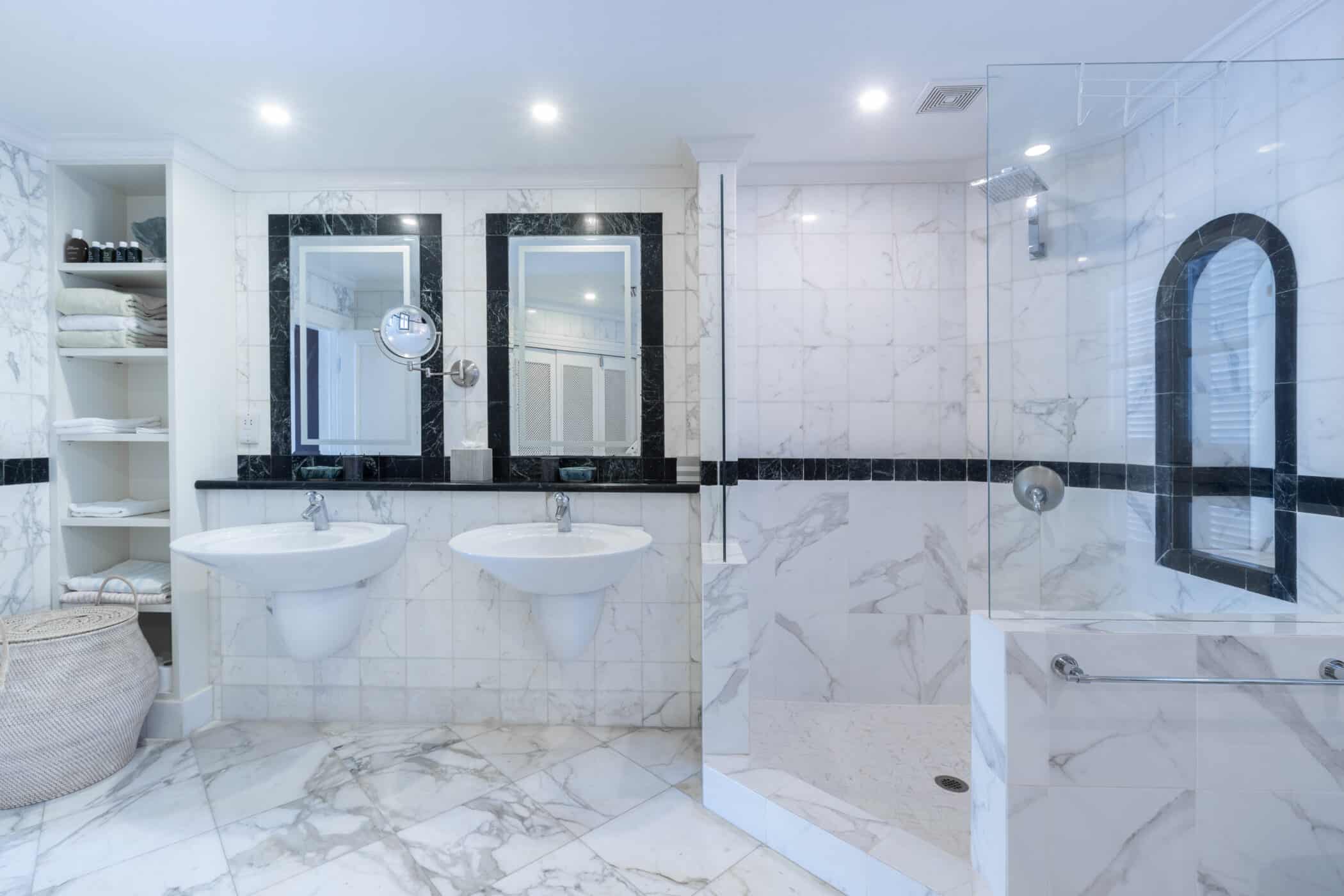 Modern bathroom with marble floors and walls, double sinks, two mirrors, a glass-enclosed shower, built-in shelving with towels, and a large basket in the corner. The space is bright with recessed lighting.
