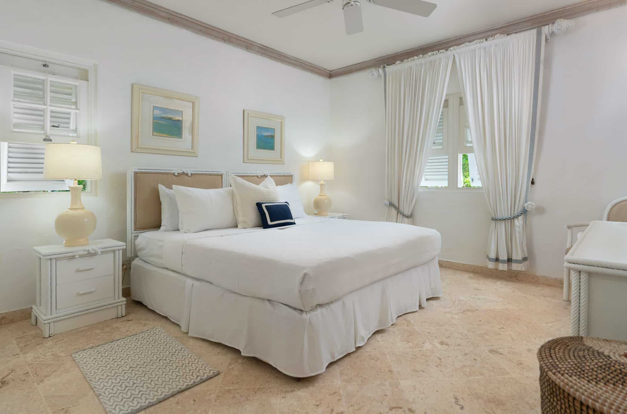 A bright, airy bedroom with a large bed, white linens, two bedside tables with lamps, framed art on the wall, white curtains, and a window letting in natural light. The floor is tiled in a light stone.