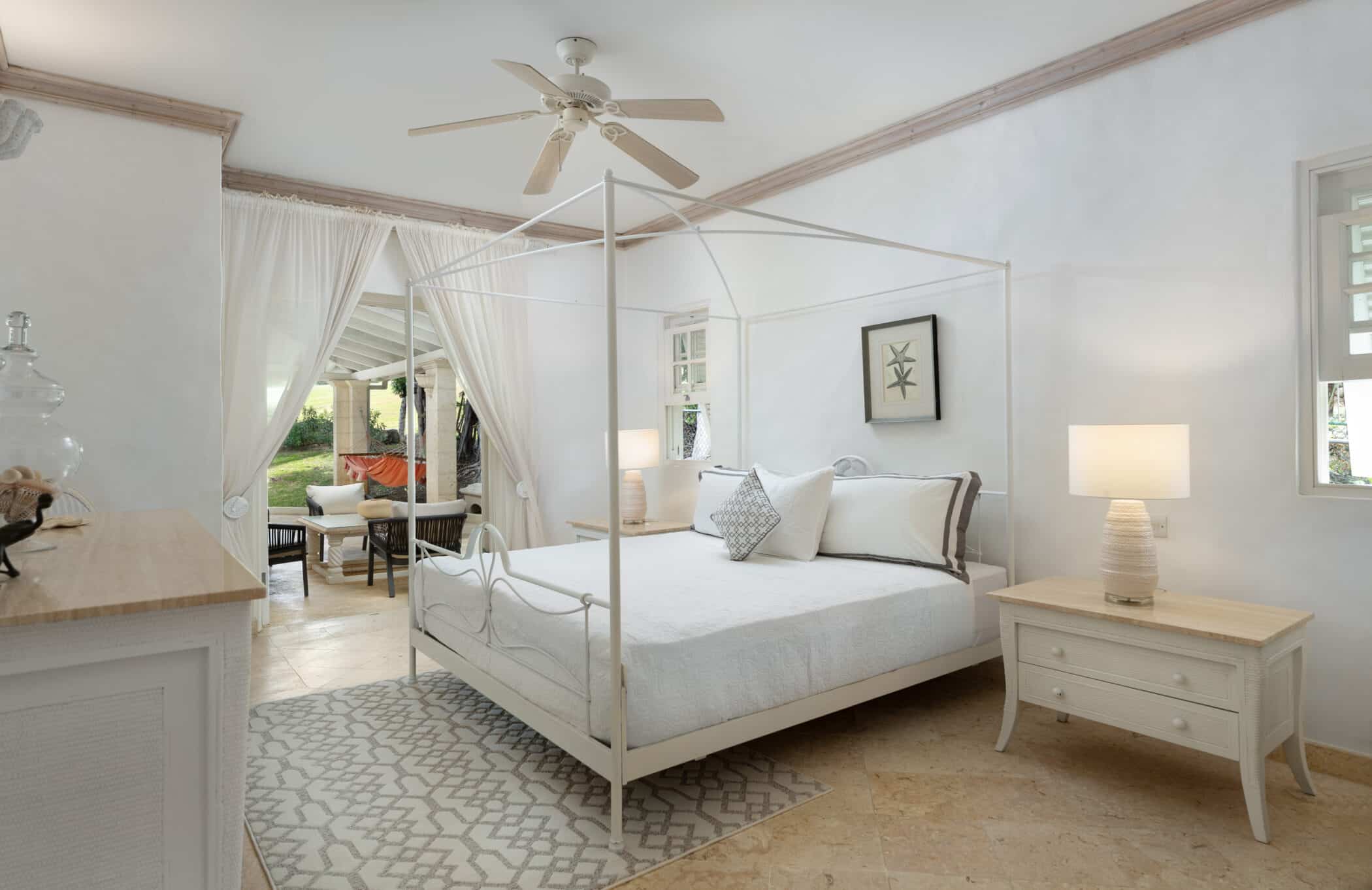 Bright, airy bedroom with a canopy bed, white linens, and neutral decor. A nightstand with a lamp sits beside the bed. Sheer curtains open to a patio with outdoor seating and greenery visible outside.