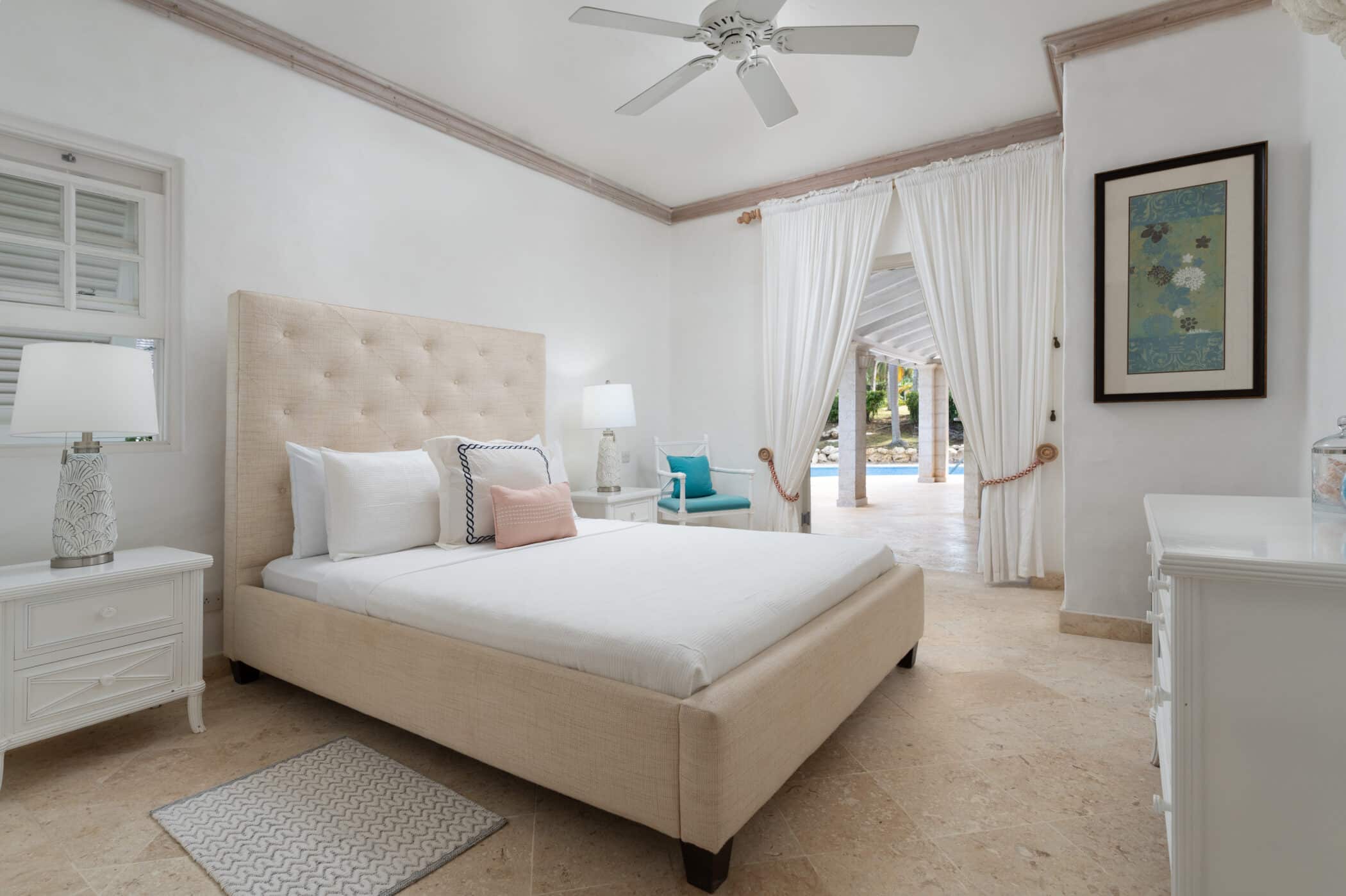 A bright, airy bedroom with a beige upholstered bed, white bedding, and bedside tables with lamps. There are white curtains, a ceiling fan, and patio doors opening to a view of a pool and greenery outside.