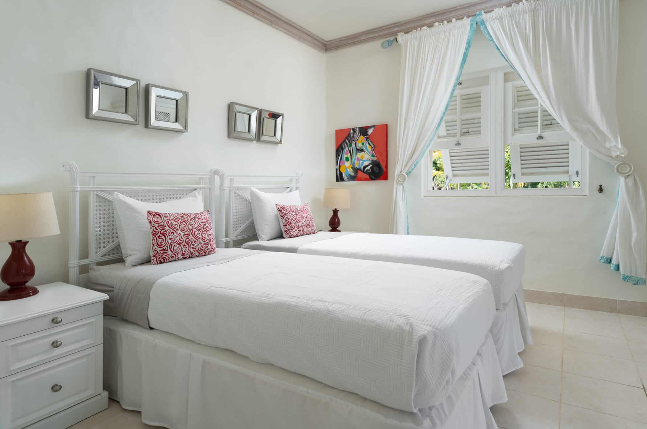 A bright bedroom with two twin beds side by side, white bedding, red accent pillows, a nightstand with a lamp, three square mirrors on one wall, colorful horse artwork, and a window with white curtains.