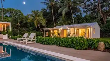 A small, modern white house with large windows is surrounded by lush greenery and palm trees, next to a pool with lounge chairs at dusk. The sky is clear with a visible full moon.