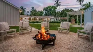 Cozy outdoor seating area with cushioned chairs arranged around a fire pit, set on a stone patio. Modern cottages and palm trees are in the background under a colorful sunset sky.