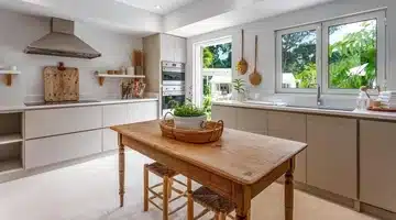 Bright, modern kitchen with light cabinets, stainless steel appliances, a wooden table with two stools, and large windows overlooking greenery. Decorative utensils and plants add a cozy touch.