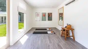 Bright yoga studio with two mats, towels, and blocks on a light wood floor. Large windows let in natural light. Art with floral prints hangs on the wall next to a large mirror and a wooden bench with yoga accessories.