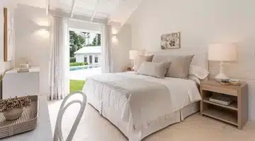 A bright, airy bedroom with white walls and bedding, light wood furniture, and two bedside lamps. French doors open to a view of a green lawn, a pool, and a white building outside.