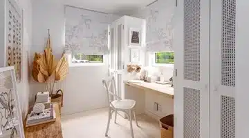 Bright, tidy home office with white walls, built-in desk, chair, two windows with patterned blinds, dried palm leaves, framed art, and woven décor accents.