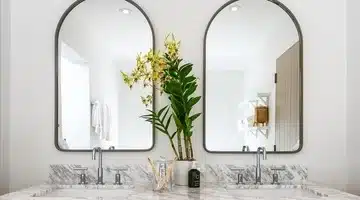 A modern bathroom features a double sink with a marble countertop. Two large arched mirrors hang above the sinks. A tall vase with green leaves and yellow flowers sits between the sinks. The walls are white.