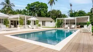 Modern outdoor swimming pool surrounded by wooden decking, white lounge chairs, umbrellas, and cabanas, with lush green trees and a clear sky in the background.