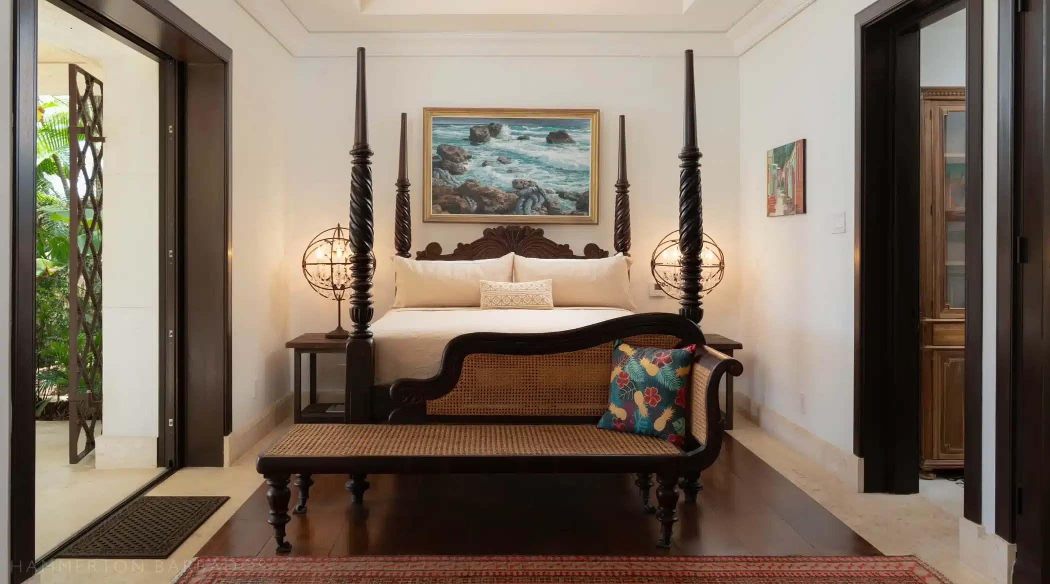 A stylish bedroom with a four-poster bed, globe lamps on each side, a wooden bench with a colorful pillow at the foot of the bed, and a seascape painting hanging above the headboard.
