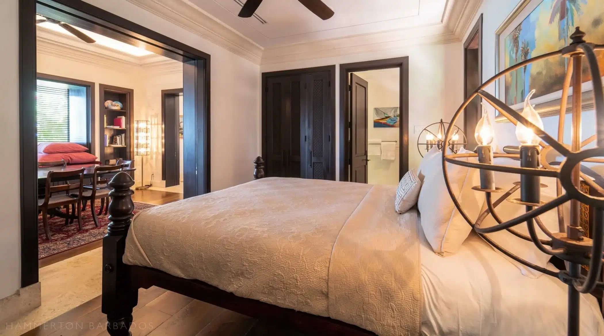A cozy bedroom with a large bed, beige bedding, and decorative pillows. There are open doorways leading to a study with bookshelves and a dining room. Warm lighting and artwork create a welcoming atmosphere.