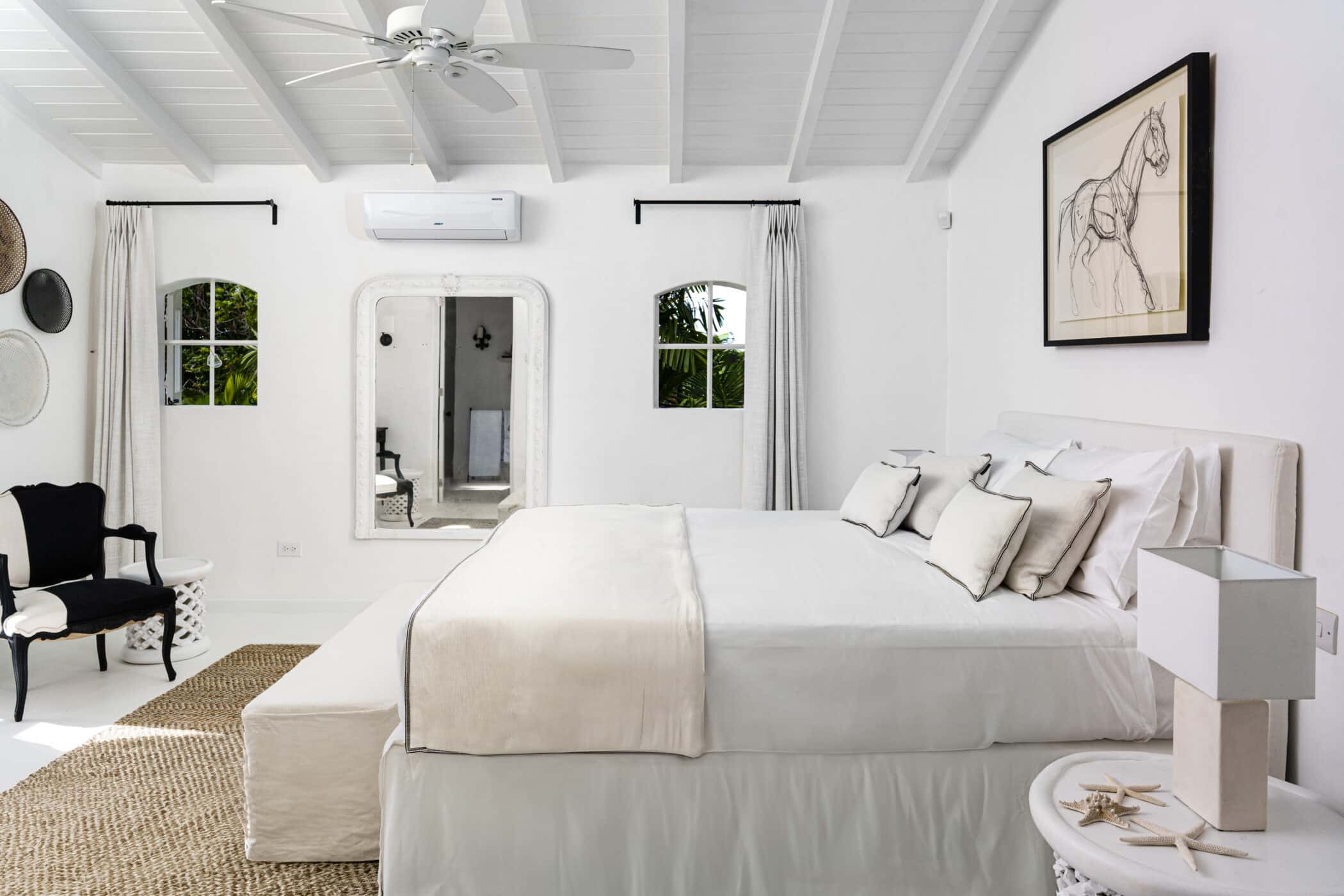 Bright, airy bedroom with white walls, bed, and ceiling. Large mirror, minimalist decor, framed horse sketch above bed, woven rug, black armchair, two curtained windows, and soft natural light create a serene, modern space.