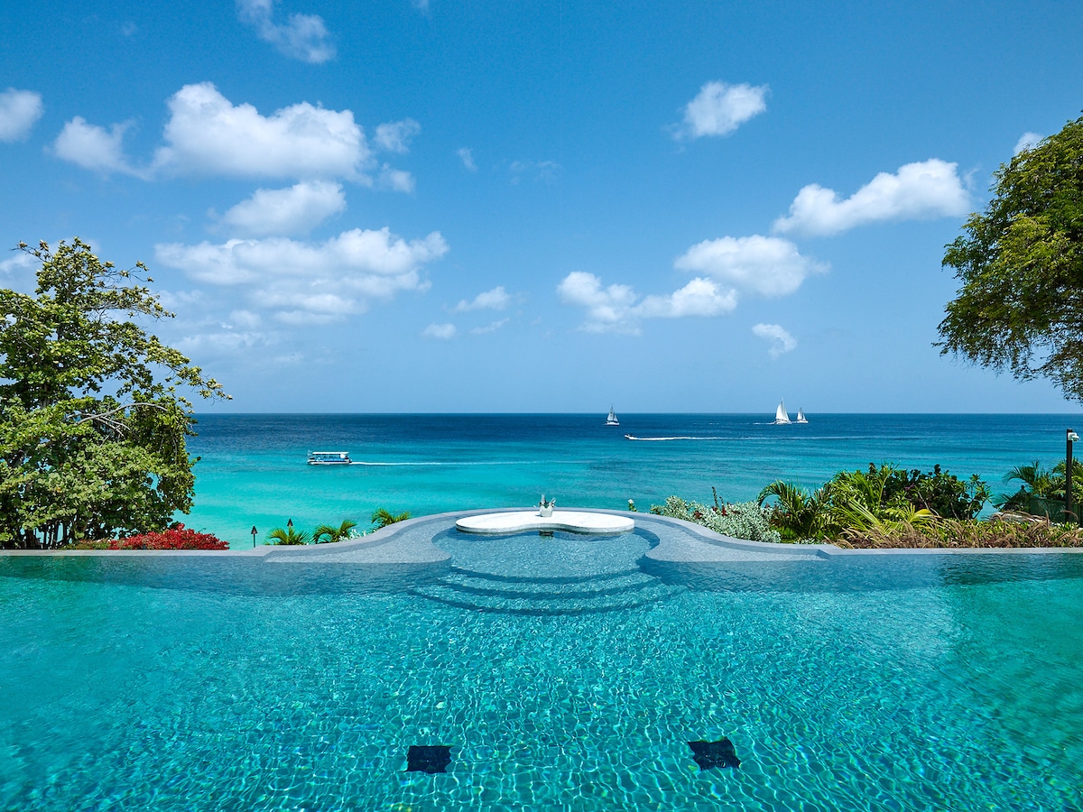 Infinity pool overlooking a turquoise ocean with sailboats and a small boat in the distance under a bright blue sky with scattered clouds, surrounded by lush tropical greenery.