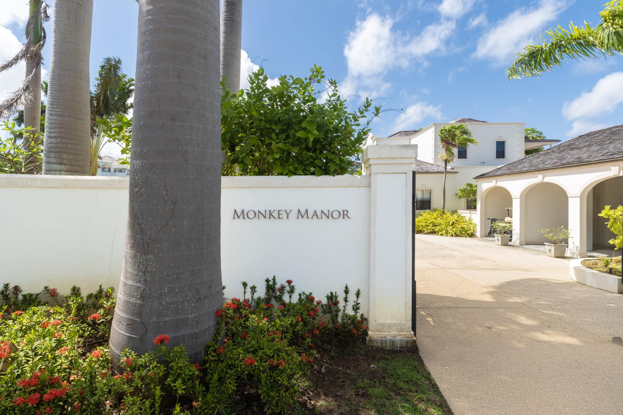 A white wall with Monkey Manor written on it stands beside tall palm trees and red flowers, with a large white house and blue sky in the background.