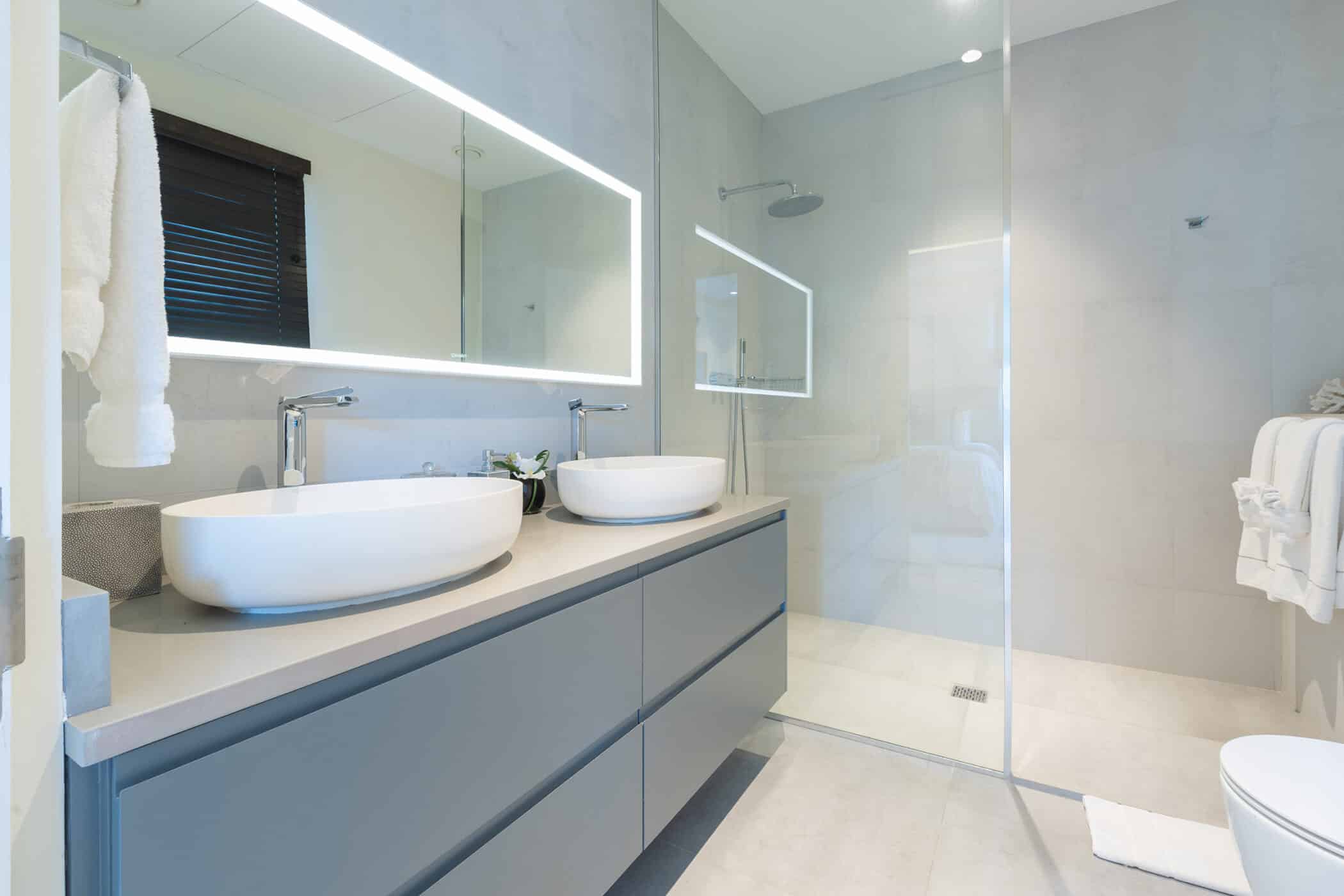 Modern bathroom with a large mirror, two white vessel sinks on a gray vanity, a glass-enclosed walk-in shower, towel racks, and neutral-toned walls and floors.