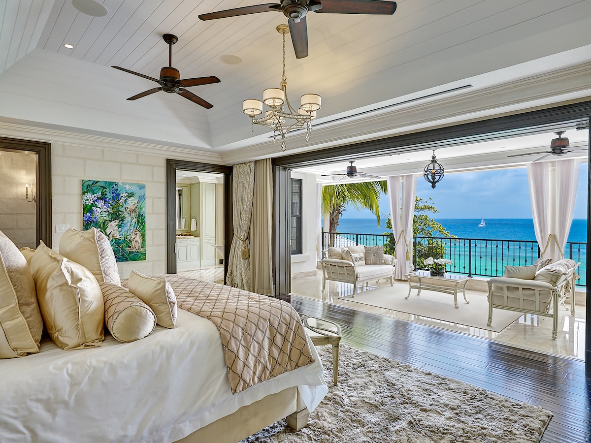 Luxurious bedroom with beige bedding, chandelier, and ceiling fans, opening to a balcony with white seating, ocean views, and blue sky; modern décor and elegant furnishings throughout.