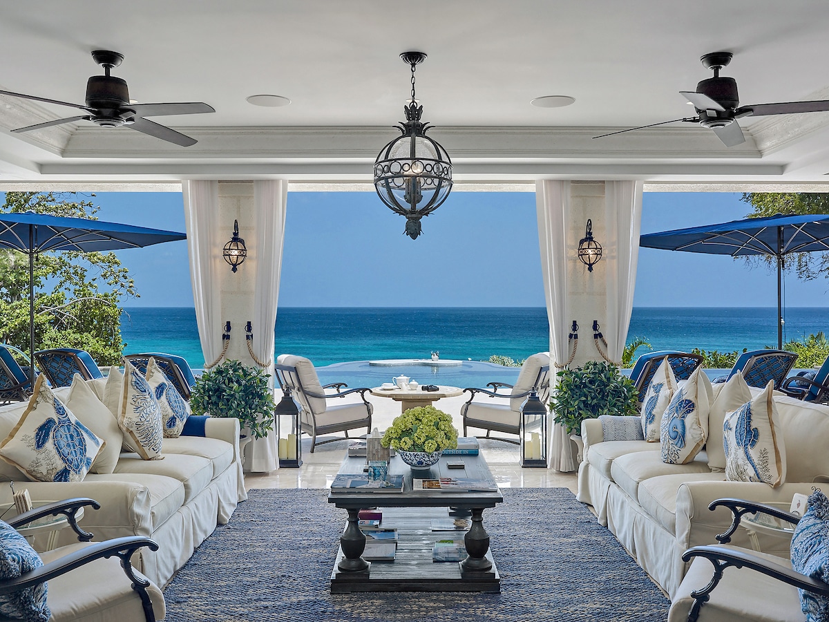 Luxurious open-air living room with white sofas, blue-patterned pillows, and a coffee table, overlooking a terrace with umbrellas and a stunning ocean view under a clear, sunny sky.