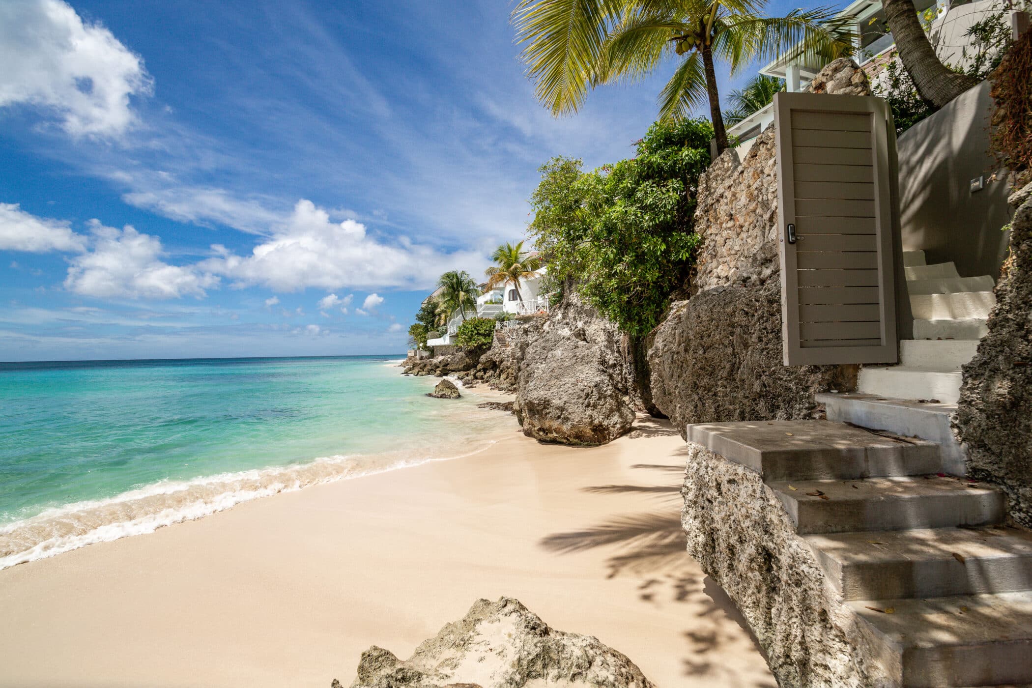 A secluded tropical beach with clear turquoise water, palm trees, and rocky cliffs. Stone steps lead down from a doorway to the sandy shore under a sunny blue sky with scattered clouds.