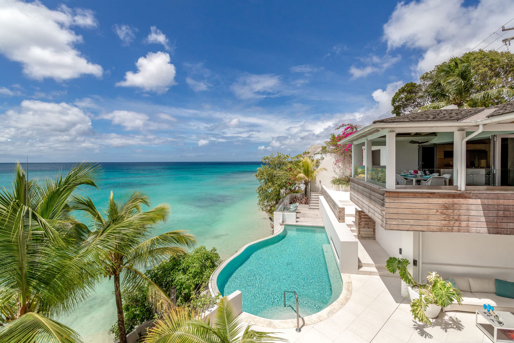 A luxurious villa with an infinity pool overlooks a turquoise ocean and sandy beach, surrounded by palm trees and lush greenery under a bright blue sky with scattered clouds.