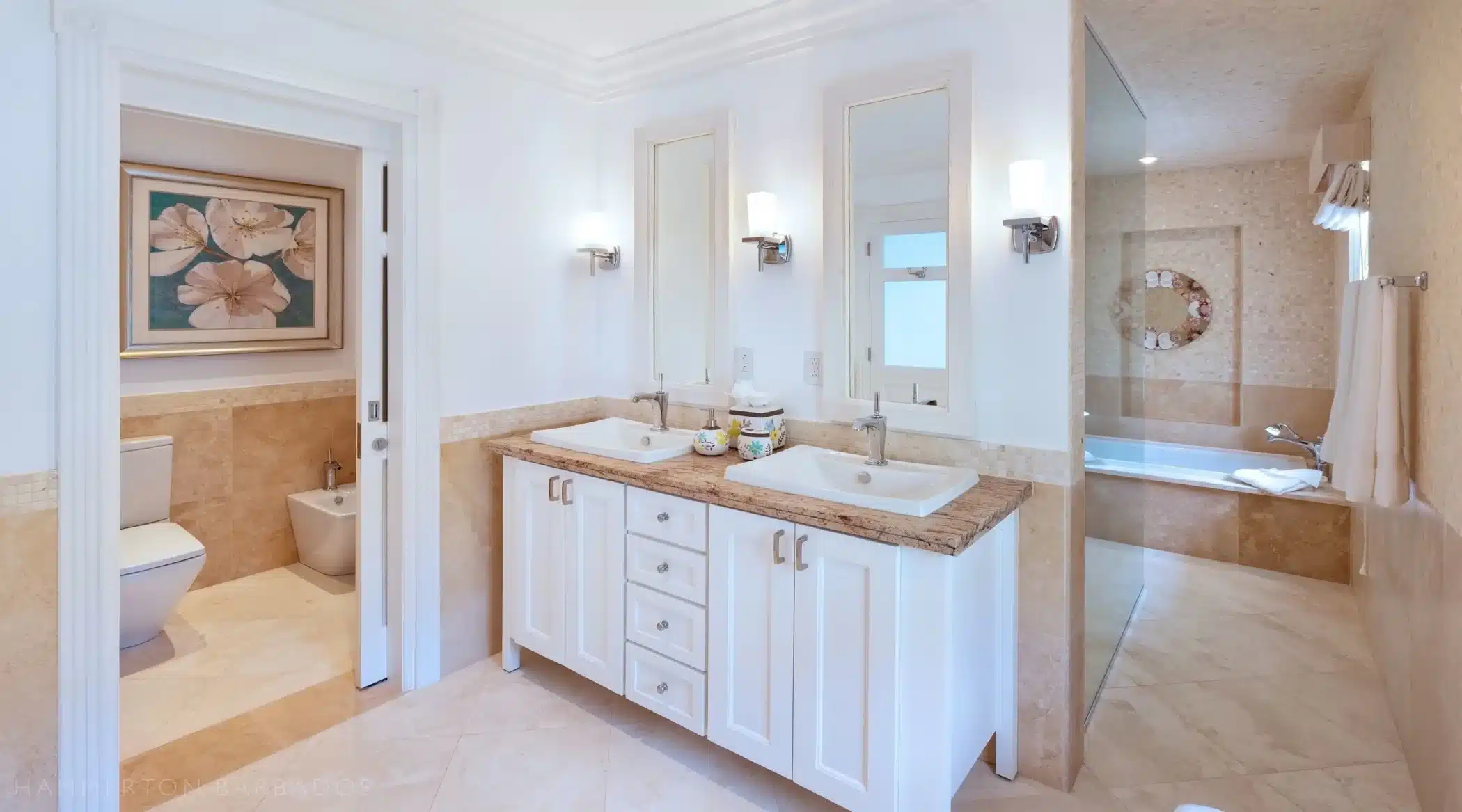 A bright, modern bathroom with double sinks on a beige countertop, white cabinetry, wall mirrors with sconces, a separate toilet area, and a spacious bathtub in a tiled alcove.
