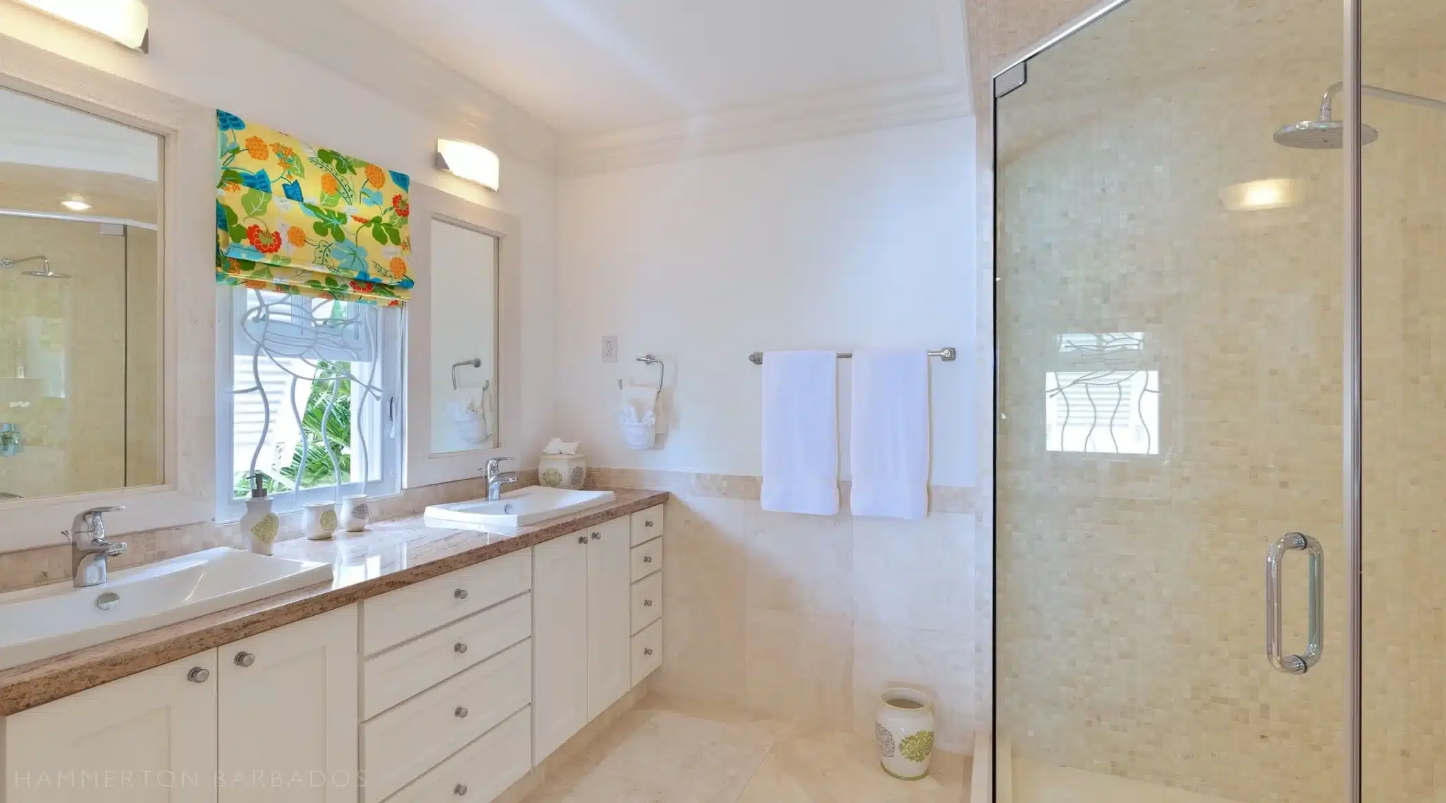 Bright bathroom with a double sink vanity, white cabinets, a large mirror, colorful floral window shade, towel rack with white towels, and a glass-enclosed shower with beige tile.