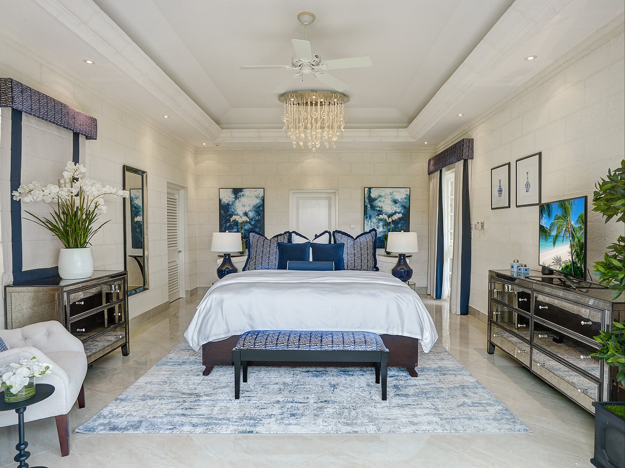 Elegant bedroom with a large bed, blue and white decor, a chandelier, two mirrored dressers, a TV, framed artwork, and plants. The room features a patterned rug and a bench at the foot of the bed.