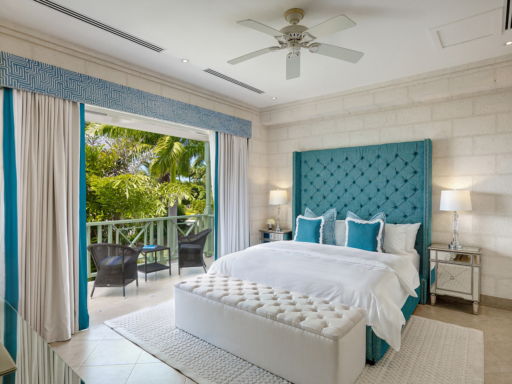 A bright, elegant bedroom with a large tufted teal headboard, white bedding, and pillows. The room opens to a balcony with two chairs, overlooking lush tropical greenery. A ceiling fan and bedside lamps complete the space.