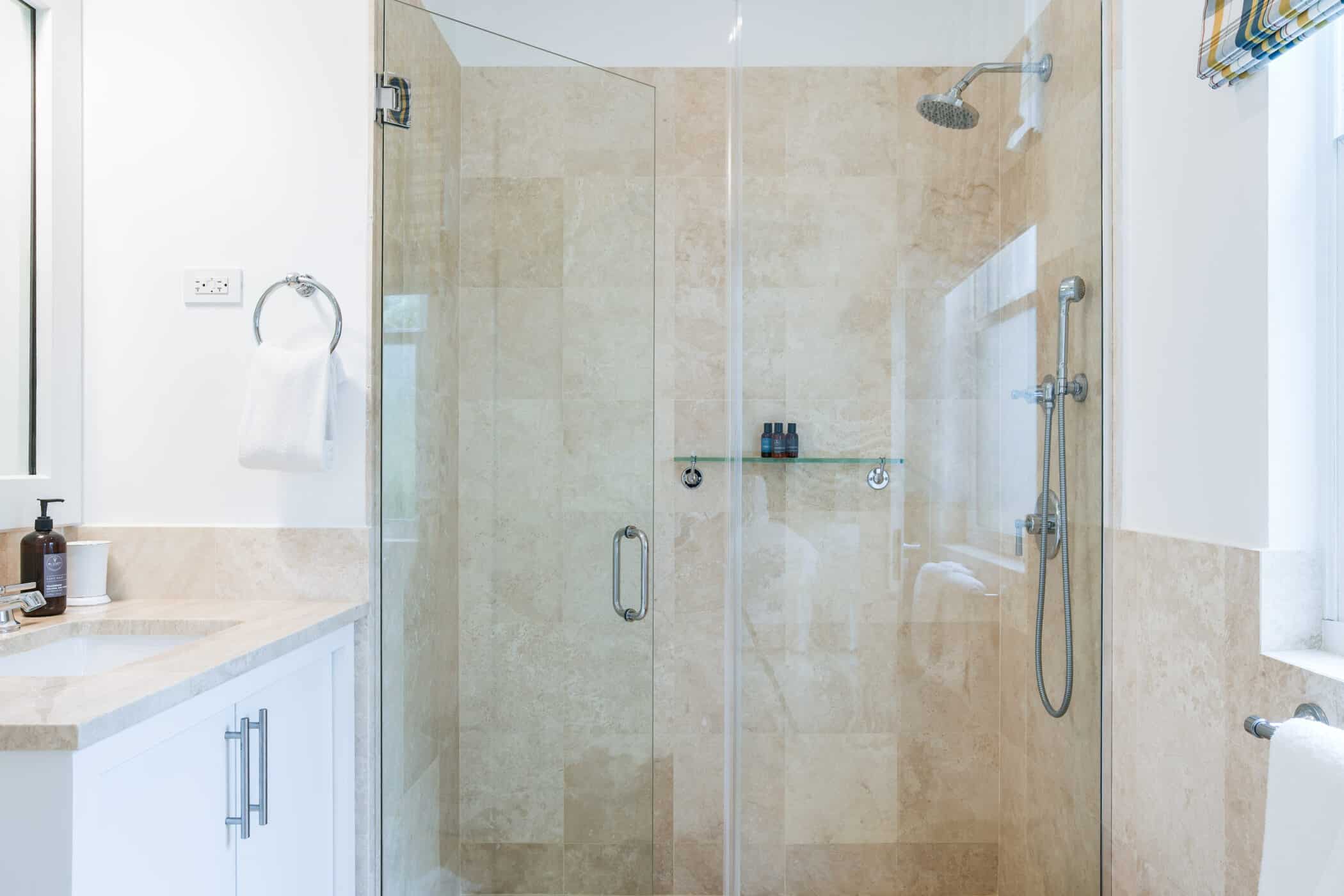 A modern bathroom with a glass-enclosed shower, beige marble walls, a wall-mounted showerhead, and a glass shelf holding toiletries. The sink counter is white with a soap dispenser and towel nearby.
