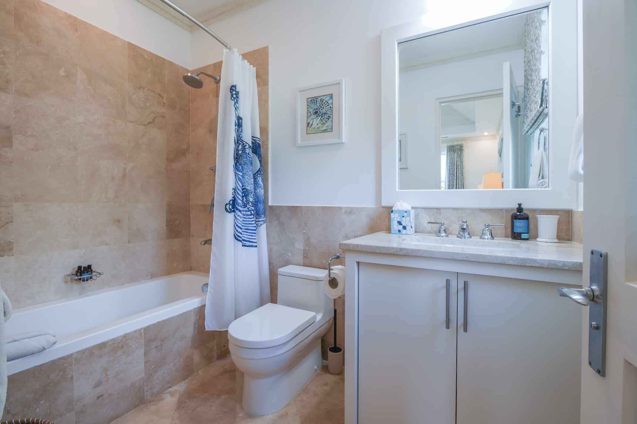 Modern bathroom with tan tiled walls and floor, a bathtub with a white and blue shower curtain, a white toilet, and a vanity with a large mirror, soap dispenser, and tissue box. A framed picture hangs on the wall.