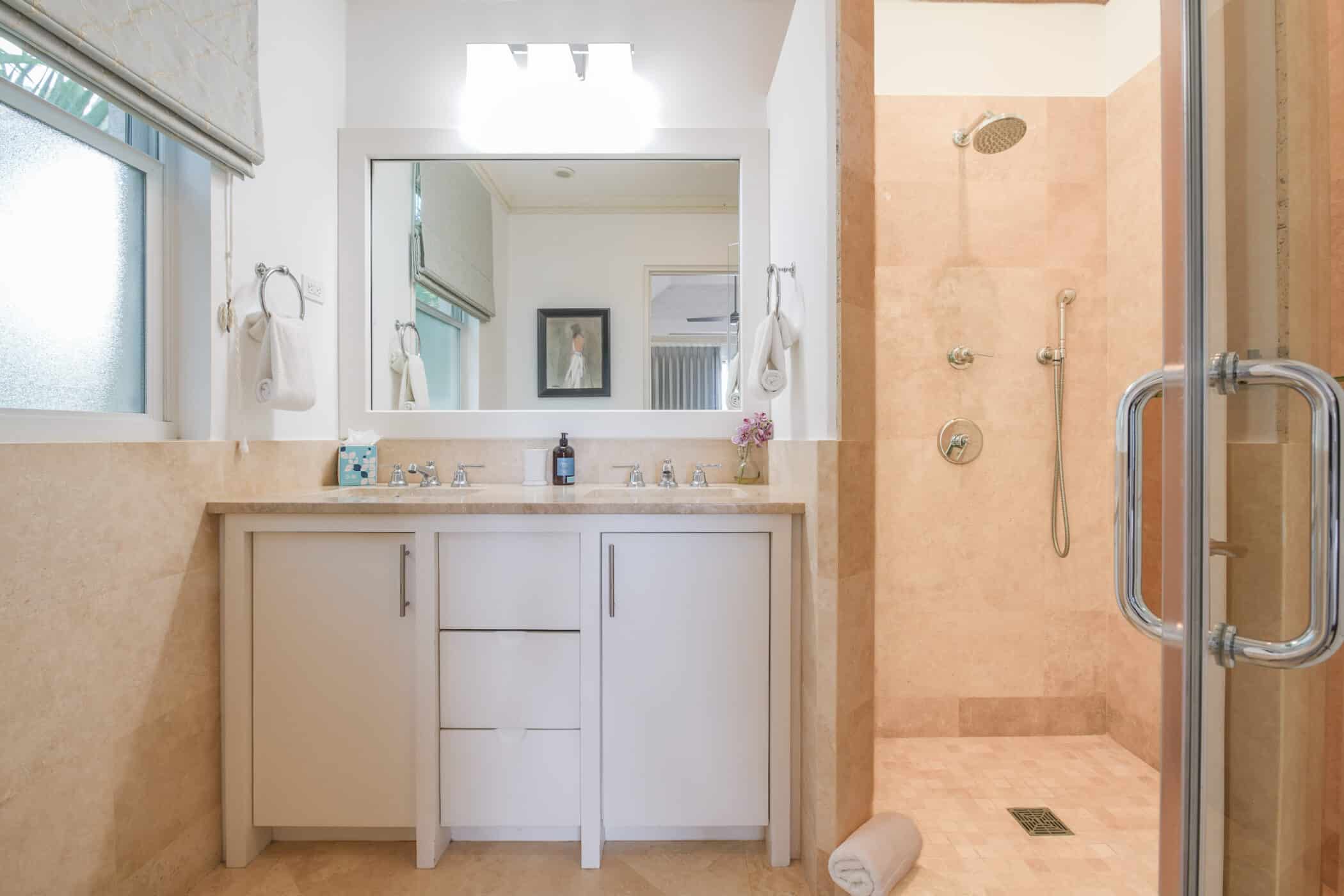 Bright bathroom with a double-sink vanity, large mirror, towels, and toiletries. On the right is a spacious walk-in shower with beige tiles and multiple showerheads. A rolled towel sits on the floor near the shower.