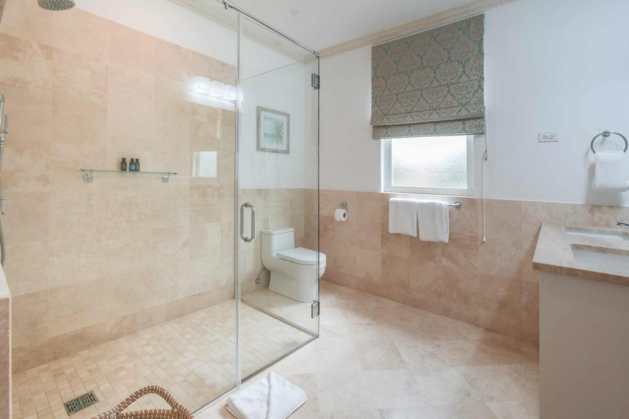 Modern bathroom with beige tile walls and floor, glass-enclosed shower, toilet, towel racks, window with patterned roman shade, and double sink countertop. Bright and clean with simple decor.