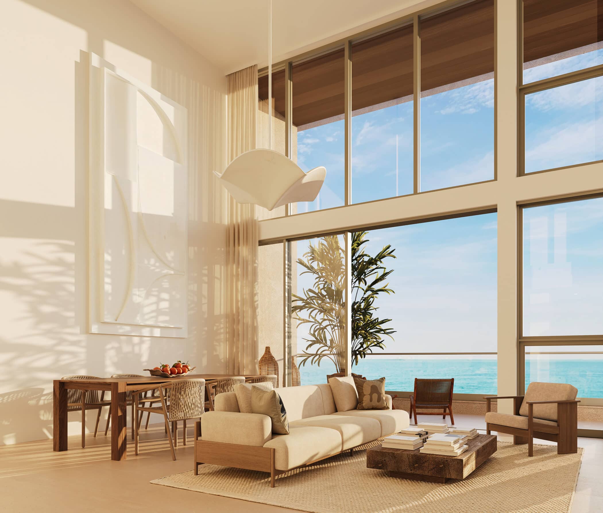 A bright, modern living room with tall windows overlooking the ocean, furnished with a beige sofa, wooden chairs, a rustic coffee table, and a dining area with a fruit bowl. Sunlight fills the airy, spacious space.