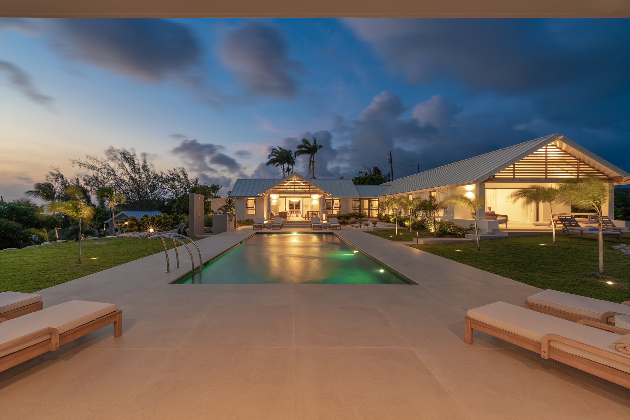 Luxury villa at sunset with a lit swimming pool, surrounded by lounge chairs, palm trees, and modern outdoor lighting, creates a serene and inviting atmosphere reminiscent of the tranquil elegance found at Cloudbreak.