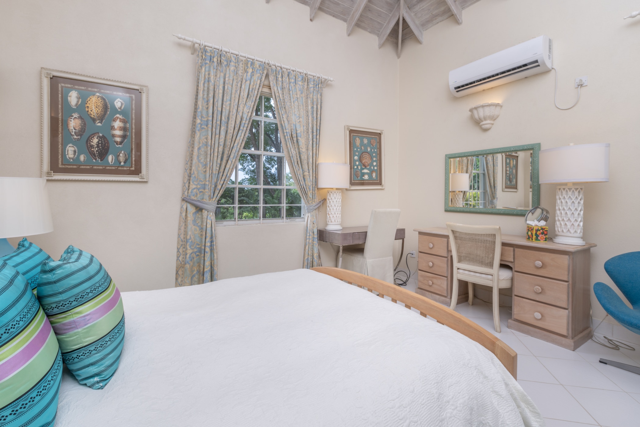 Bright bedroom with a white bed, colorful pillows, a wooden desk with a mirror, and a window with patterned curtains. Wall art, lamps, and an air conditioner decorate the cream-colored walls.