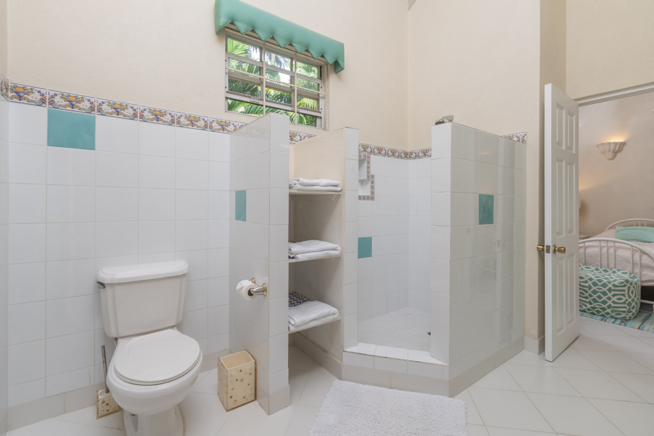 A bright bathroom with a white toilet, open shelves with towels, a walk-in shower with blue and white tiles, and a door leading to a bedroom with a bed and turquoise bedding.