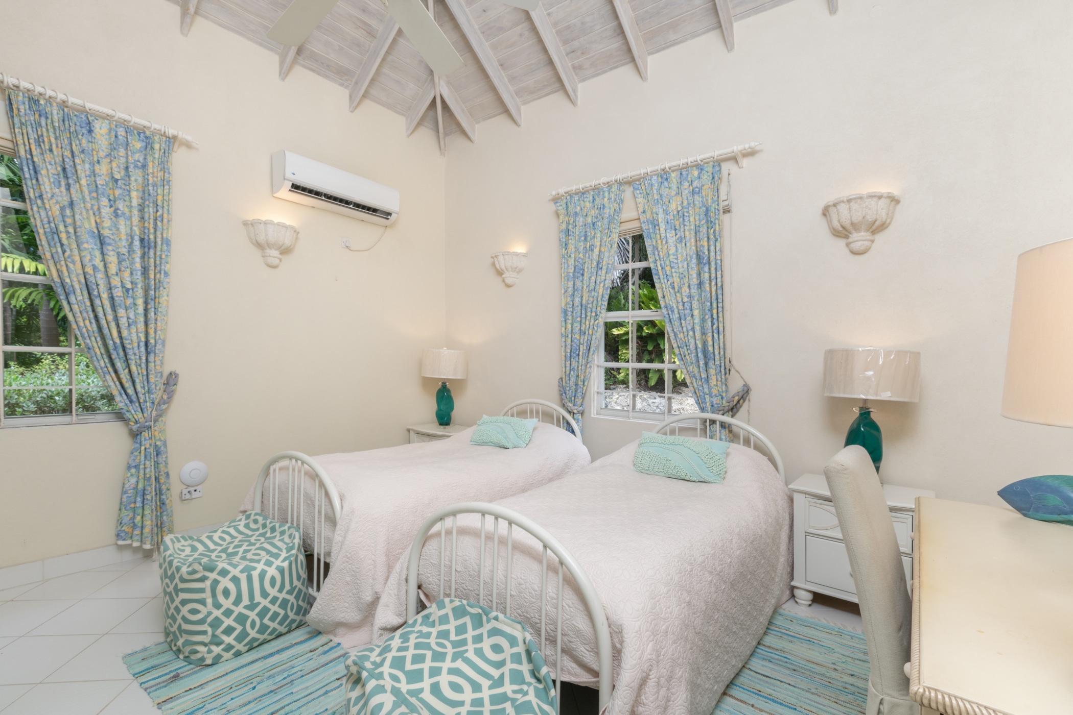 A bright bedroom with two twin beds, teal and white patterned stools and curtains, light bedding, a desk with a lamp, air conditioner, and windows showing green trees outside. The ceiling is sloped with exposed beams.