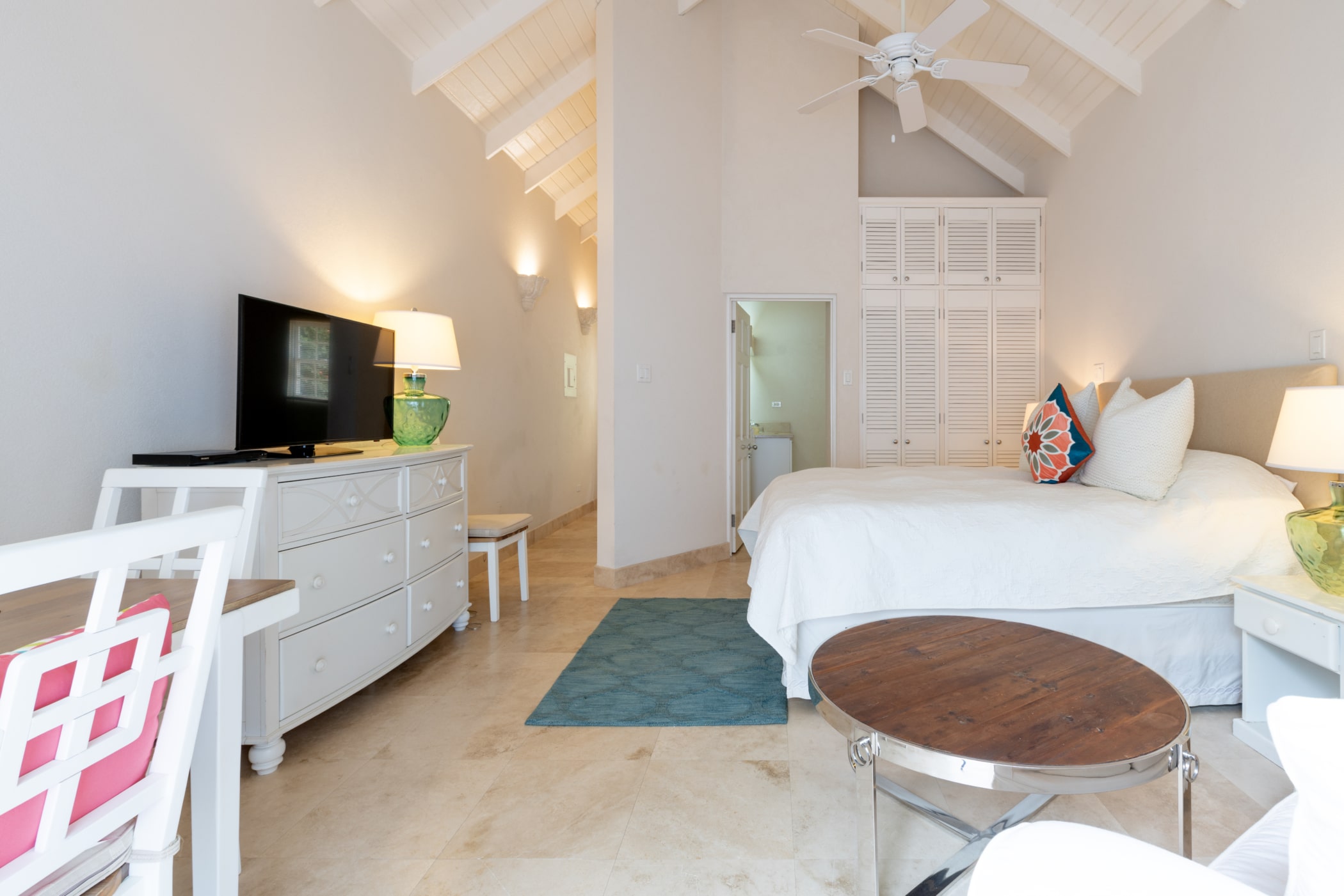 A bright, airy bedroom with a white vaulted ceiling, double bed with colorful pillows, a TV on a white dresser, a round wooden coffee table, and a small desk and chair. Natural light fills the room.
