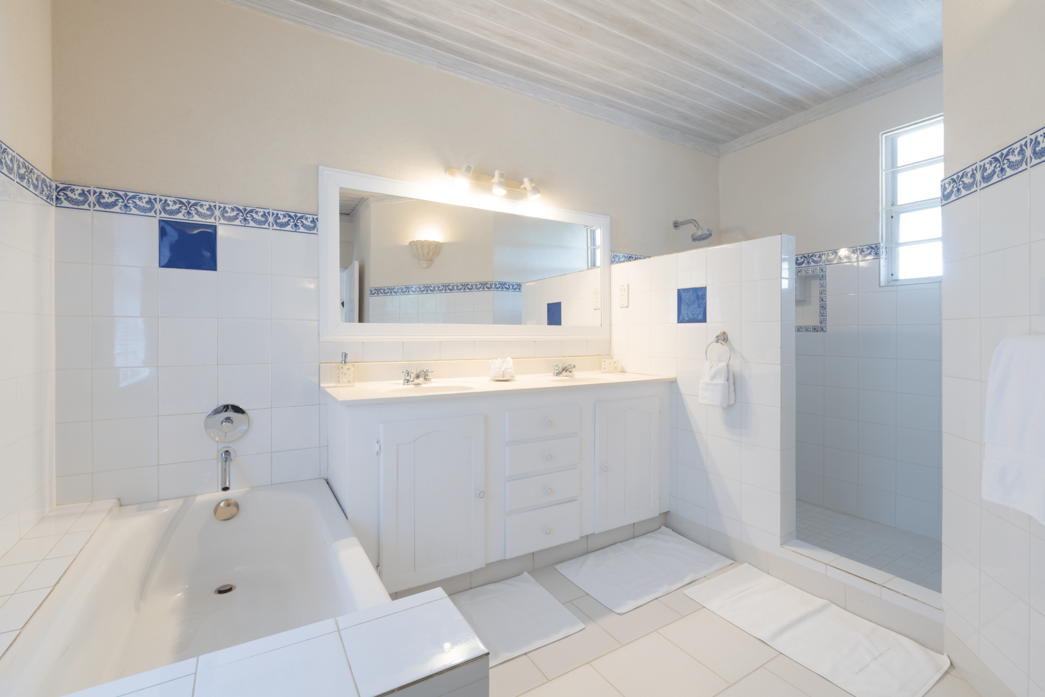 A bright, white-tiled bathroom with a bathtub, double sink vanity, large mirror, walk-in shower, and blue decorative tile accents. White bath mats are placed on the floor. A small window lets in natural light.