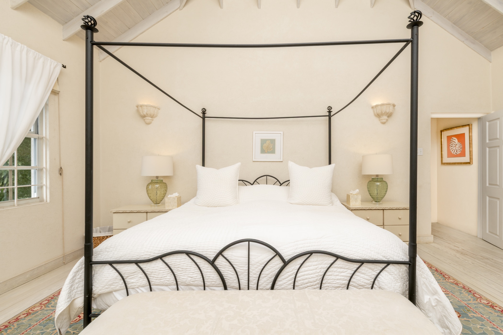 Bright bedroom with a black metal four-poster bed, white bedding, two green glass lamps on bedside tables, a framed picture above the bed, and white walls with large windows and high ceilings.