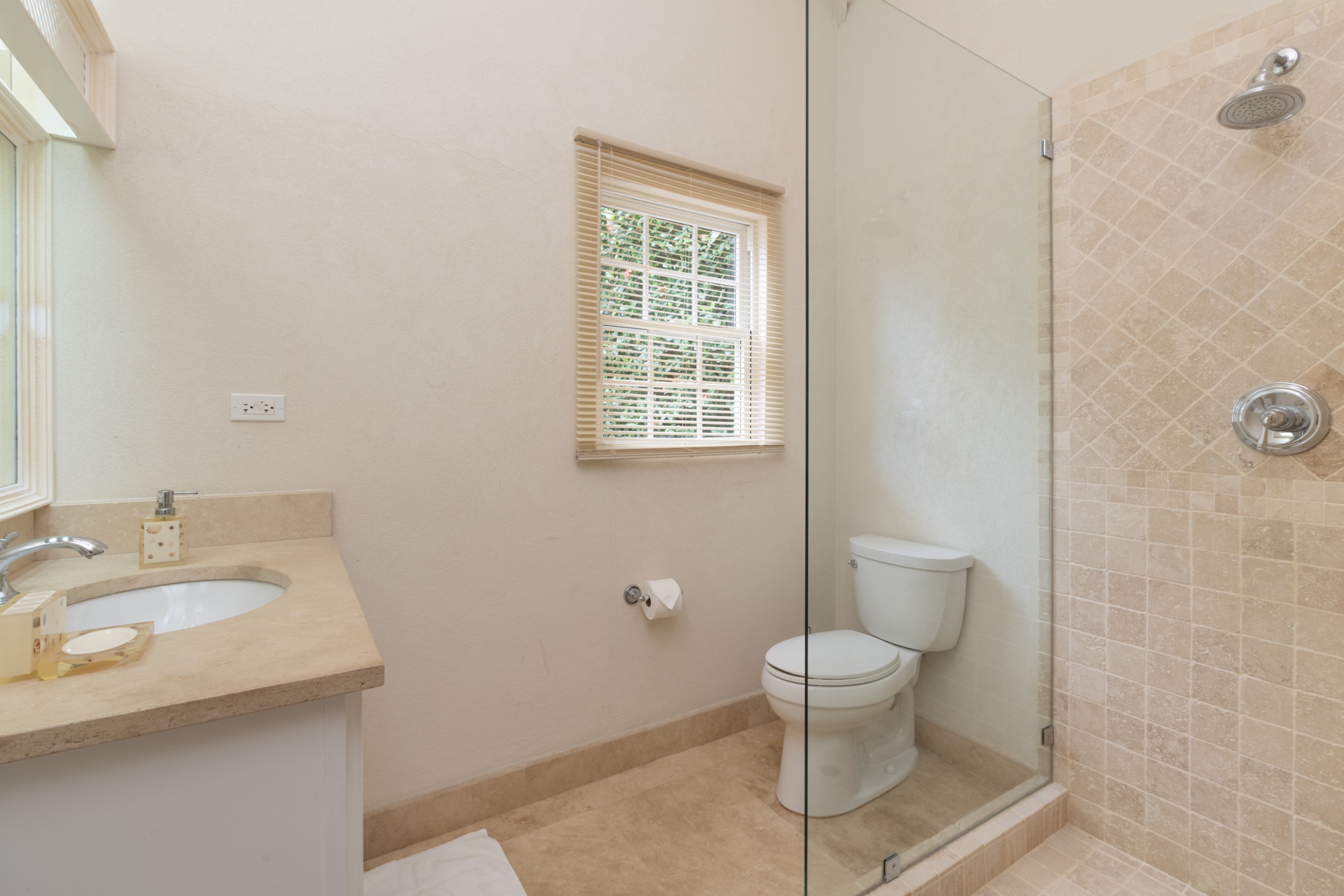 A clean bathroom with a beige countertop sink on the left, a toilet in the center, and a glass-enclosed shower with tan tile walls on the right. A window with blinds lets in natural light.